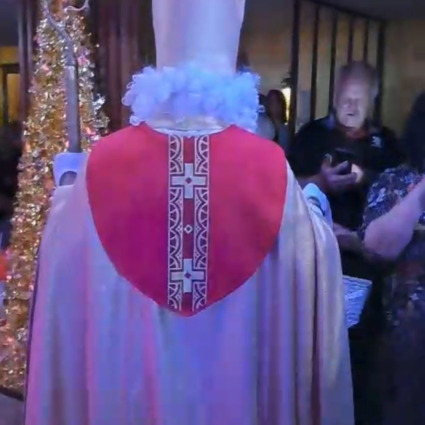 Ein Priester steht in einer Kirche, den Rücken zugewandt, und trägt eine rot-weiße Veste. Hinter ihm steht ein goldener Weihnachtsbaum, und eine Menschenmenge, einschließlich eines älteren Mannes und einer Frau, ist anwesend.