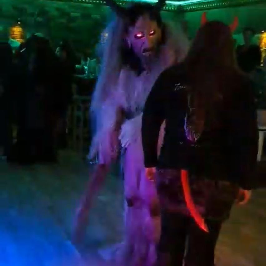 Eine Person in einem Dämonenkostüm mit roten Augen und Hörnern steht in einem schwach beleuchteten Raum mit lila Lichtern. Eine andere Person steht neben ihr in einem schwarzen Outfit.