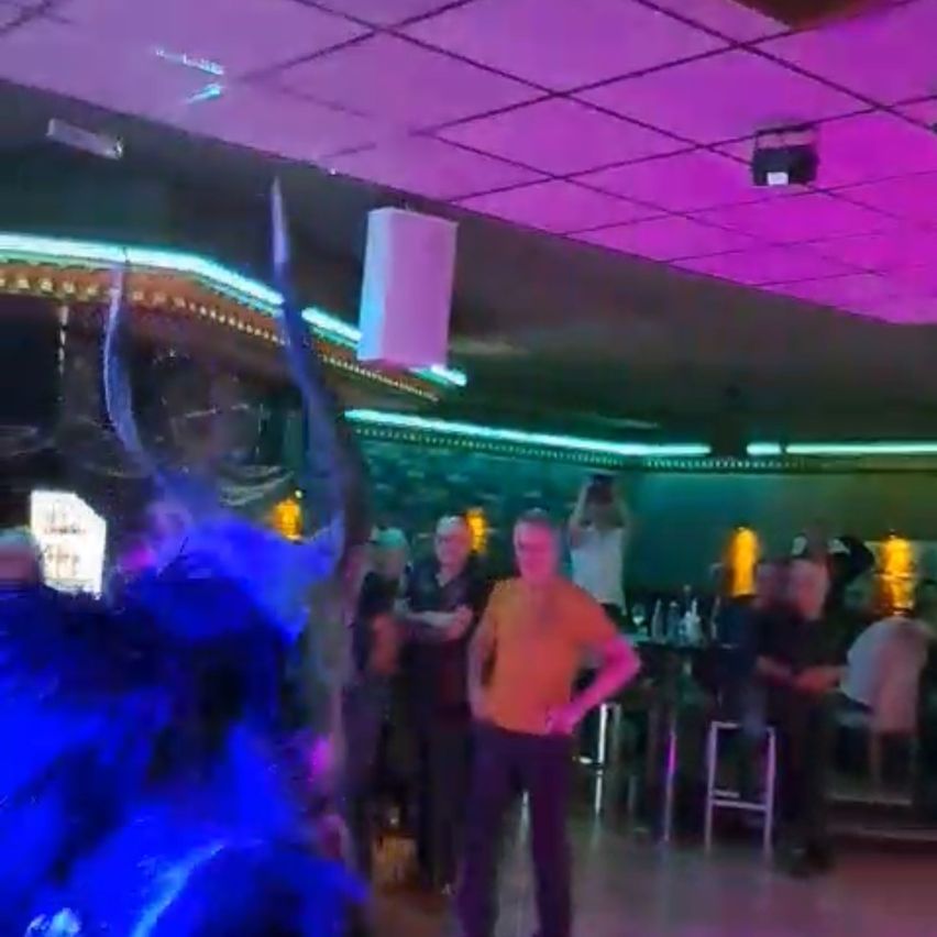 Ein Mann in einem orangefarbenen T-Shirt tanzt in einem Club mit lila Beleuchtung. Hinter ihm sitzen Menschen auf Stühlen und schauen zu.