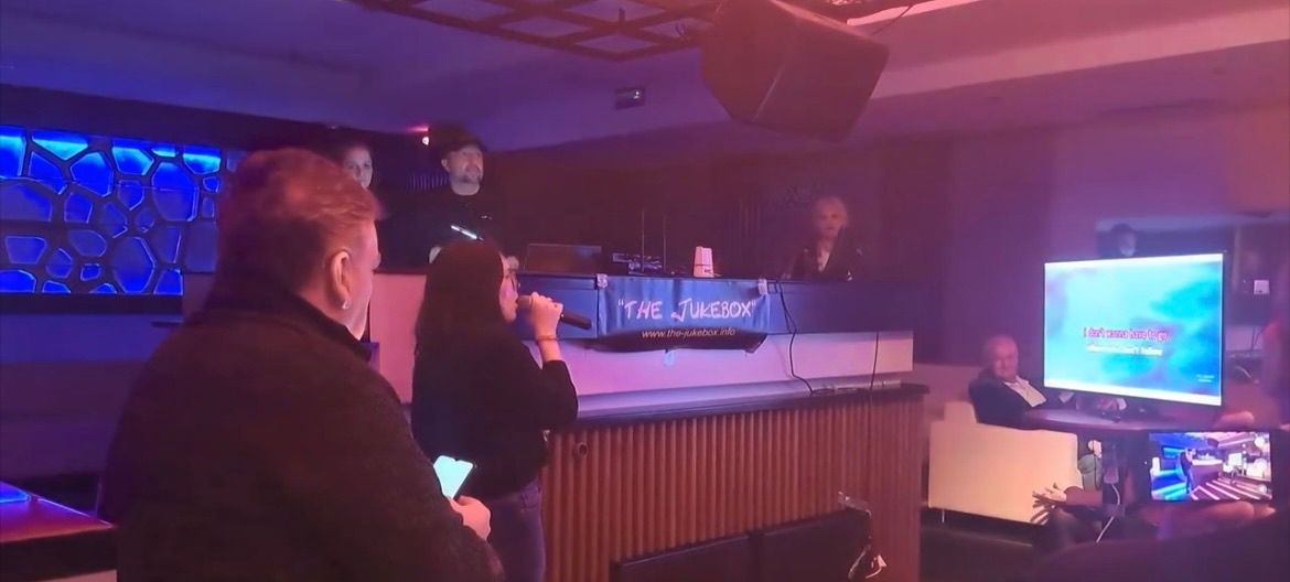 Eine Frau singt in ein Mikrofon bei einer Karaoke-Veranstaltung, mit einem Mann neben ihr. Dahinter sitzt ein DJ an einer Kabine mit einem Schild, das 'The Jukebox' steht. Mehrere Personen sitzen im Hintergrund, und ein Monitor befindet sich auf dem Schreibtisch.
