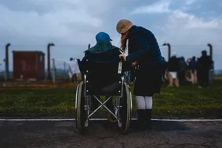 Eine Person im Rollstuhl wird von einer anderen Person in einem blauen Hut unterstützt. Sie befinden sich am Straßenrand, mit einem Zaun und einem Gebäude im Hintergrund.