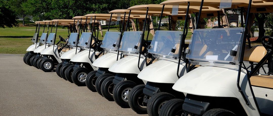 Eine Reihe weißer Golfwagen mit beigen Dächern ist auf dem asphaltierten Weg geparkt. Bäume und ein Gebäude sind im Hintergrund.