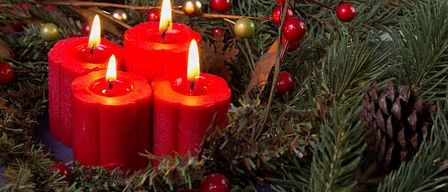 Vier rote Kerzen sind von Tannennadeln, Tannenzapfen und roten Beeren umgeben. Die Kerzen sind angezündet und werfen ein warmes Licht auf das Grün.