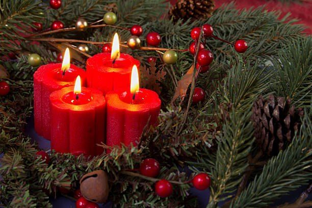 Vier rote Kerzen sind von Tannennadeln, Tannenzapfen und roten Beeren umgeben. Die Kerzen sind angezündet und werfen ein warmes Licht auf das Grün.