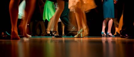 Eine Gruppe von Menschen tanzt auf einer hölzernen Tanzfläche. Einige tragen hohe Absätze. Der Hintergrund ist verschwommen.
