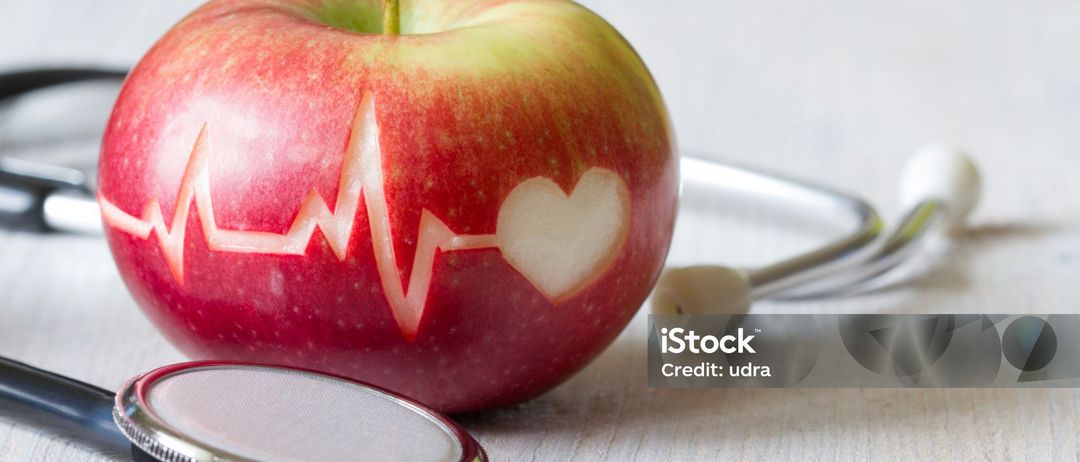 Ein roter Apfel mit einem Herz und einer EKG-Linie darauf. Daneben befindet sich ein Stethoskop. Credit: udra.