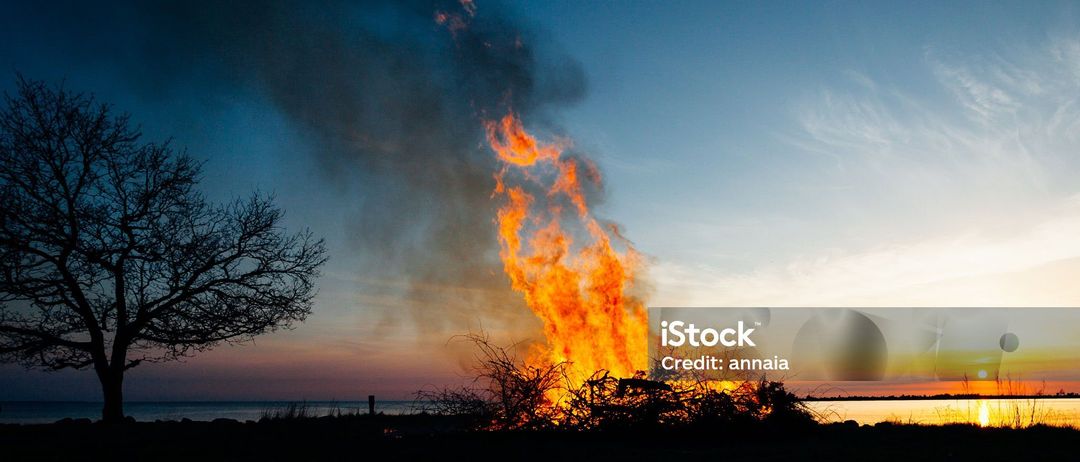 Ein Lagerfeuer brennt hell in der Dämmerung in der Nähe des Meeres, wobei Rauch in einen klaren Himmel aufsteigt.