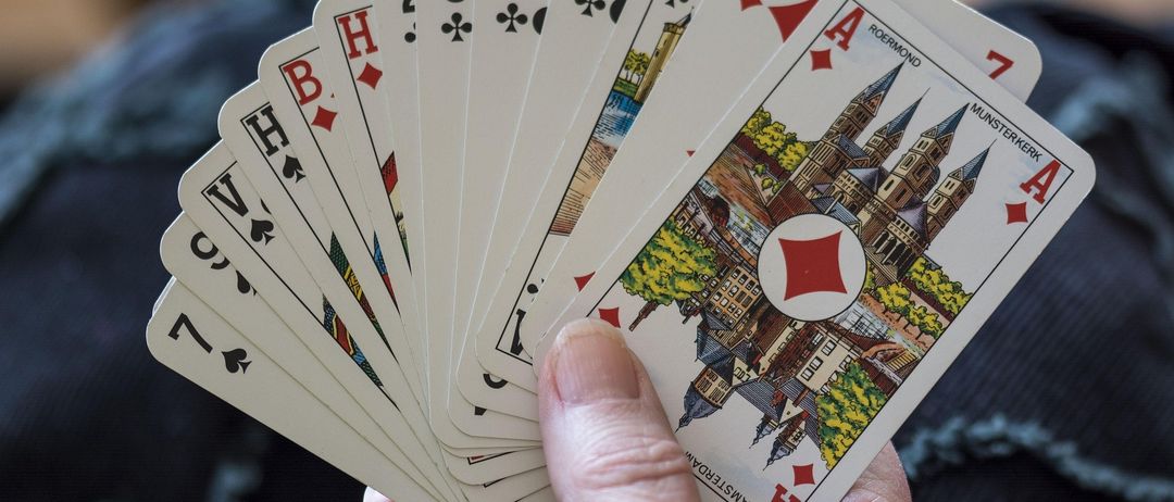 Eine Hand hält mehrere Spielkarten, darunter Ass, 7, 6, 4, 3 und einige andere, wobei das Ass der Pik im Vordergrund deutlich sichtbar ist.