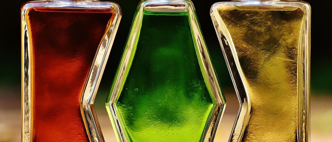 Drei Glasflaschen mit Korkenverschlüssen sind aufgereiht, jede mit einer Flüssigkeit gefüllt. Die linke Flasche enthält eine tiefrote Flüssigkeit, die mittlere eine hellgrüne Flüssigkeit und die rechte eine goldene Flüssigkeit.