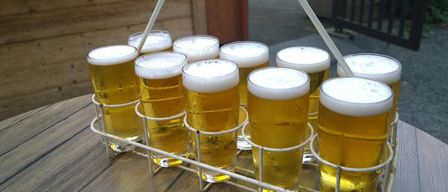 A white holder with a spiral handle carries 10 glasses of beer on a wooden table. Behind it, there is a wooden wall and a table with benches.