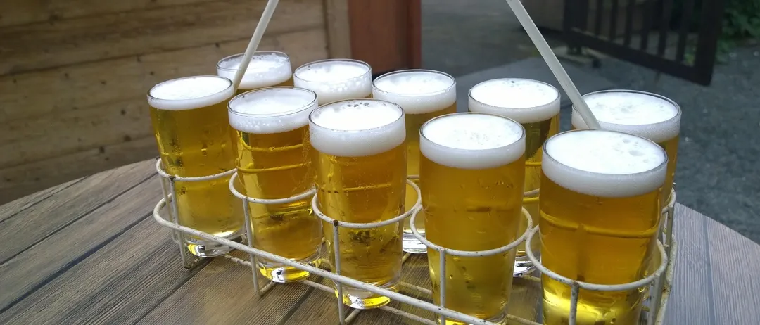 Ein weißer Halter mit Spiralgriff trägt 10 Biergläser auf einem Holztisch. Dahinter befindet sich eine Holzwand und ein Tisch mit Bänken.