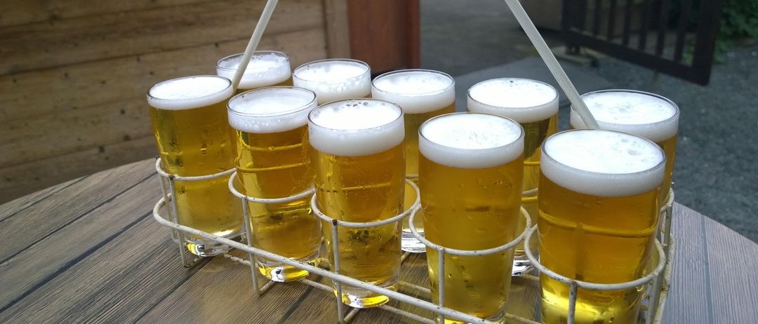 Ein weißer Halter mit Spiralgriff trägt 10 Biergläser auf einem Holztisch. Dahinter befindet sich eine Holzwand und ein Tisch mit Bänken.