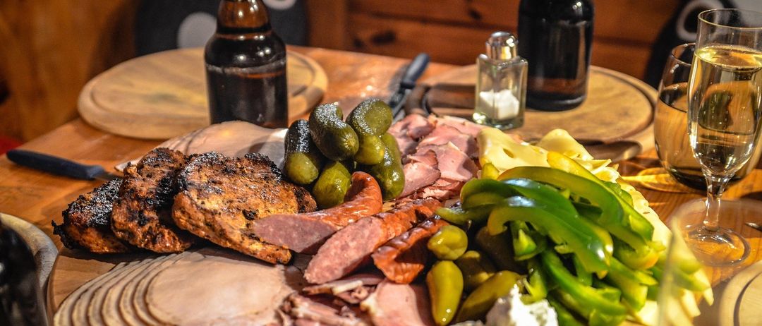 Auf einem Holztisch sind verschiedene Fleischsorten, Gurken und Käse zu sehen. Zwei Bierflaschen und eine Pfeffermühle sind ebenfalls vorhanden.