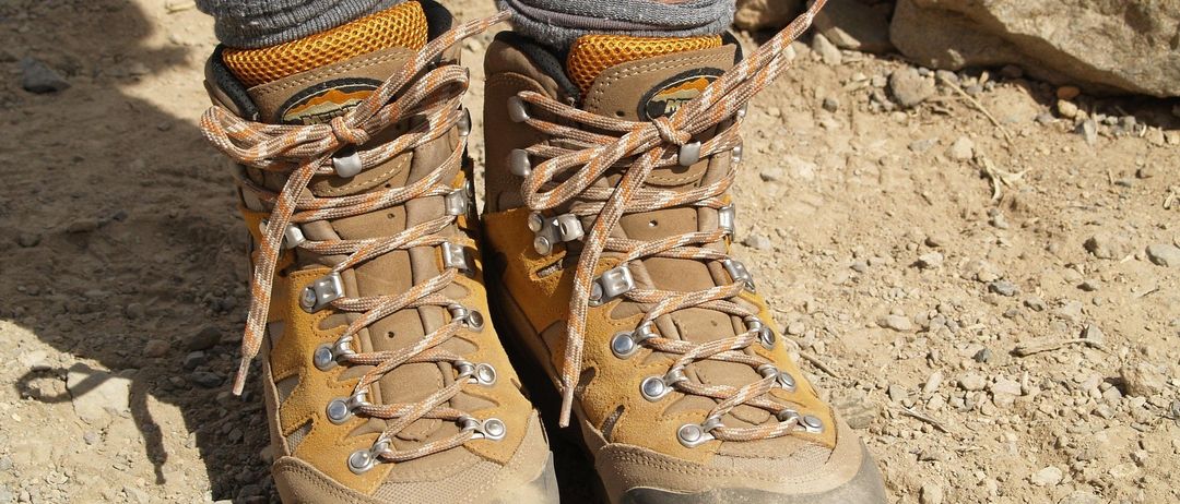 Die Beine einer Person mit Wanderschuhen, die Schnürsenkel und Riemen sowie verschmutzte Sohlen zeigen. Die Stiefel haben ein braunes und gelbes Farbschema.