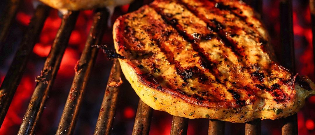 Ein Nahaufnahme von einem gut gegrillten Schweinekotelett auf einem Grill, mit sichtbaren Grillspuren und einer goldbraunen Kruste, begleitet von einem teilweise gekochten Schweinekotelett.