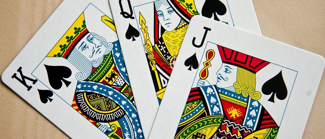 Drei Spielkarten sind zu sehen: der König der Pik, die Königin der Pik und der Bube der Pik. Sie liegen auf einer braunen Oberfläche.