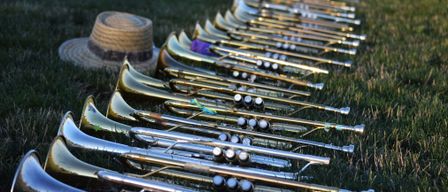 A row of trumpets are placed on grass. A straw hat is nearby. A black hat and a bottle are behind the trumpets.