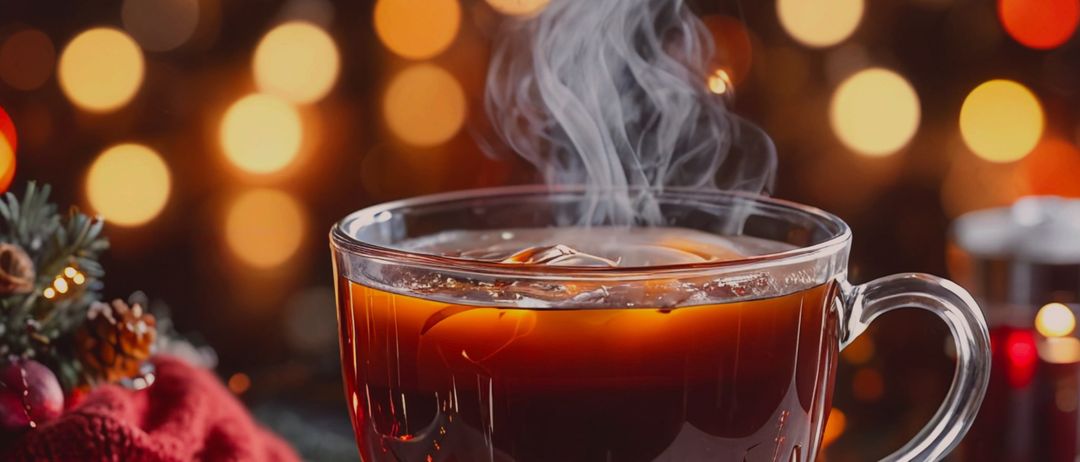 A steaming cup of tea with a red cloth and pine cones on a saucer, set against a festive background with bokeh lights.
