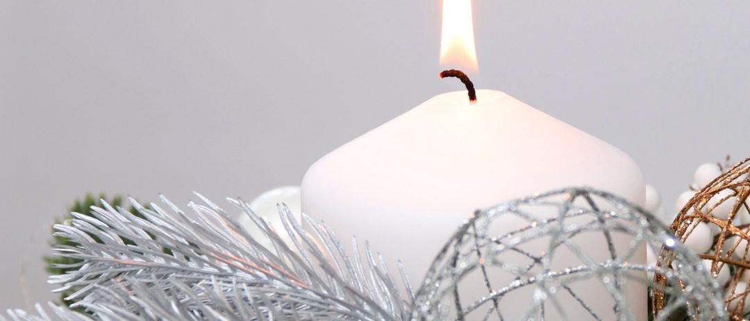 A white candle with a flame is surrounded by silver Christmas decorations, including a silver ball and pine branches.