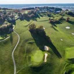Ein Luftbild eines Golfplatzes mit Häusern und Bäumen in der Ferne. Der Platz hat mehrere Löcher und die Fairways sind gut gepflegt.