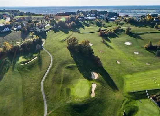 Ein Luftbild eines Golfplatzes mit Häusern und Bäumen in der Ferne. Der Platz hat mehrere Löcher und die Fairways sind gut gepflegt.