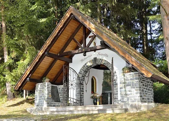 Eine kleine Kapelle mit einem Holzdach und Steinmauern befindet sich im Wald. Sie hat einen gewölbten Eingang und ein Kreuz auf der Spitze.