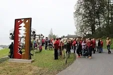 Eine Menschenmenge versammelt sich um eine große rote und braune Skulptur in einem grasbewachsenen Bereich nahe einer Straße, mit Bäumen im Hintergrund.