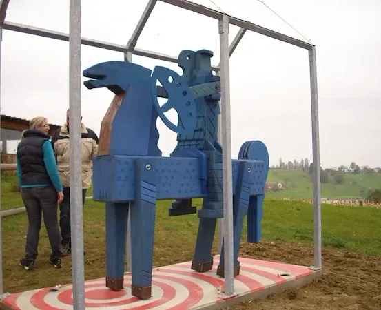 Eine blaue Holzpferdestatue steht auf einem offenen Feld, umgeben von einem Metallrahmen. Zwei Menschen in Jacken stehen in der Nähe.
