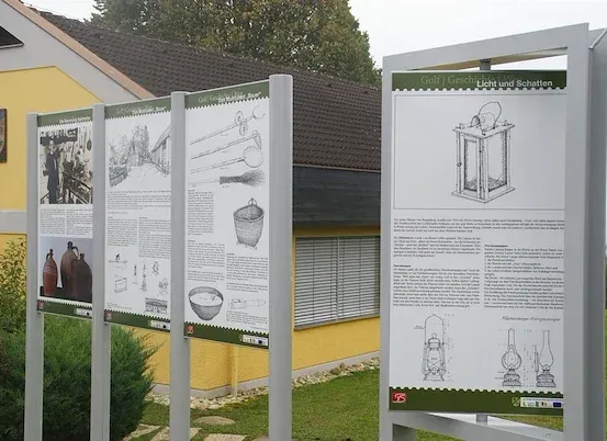 Drei Informationsschilder stehen vor einem gelben Gebäude mit einem braunen Dach. Die Schilder haben Zeichnungen verschiedener Objekte.
