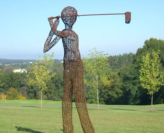 Eine große Skulptur eines Golfspielers steht auf einem Feld mit grünem Gras und Bäumen im Hintergrund. Der Golfspieler ist aus gewebtem Draht und hält einen Golfschläger.