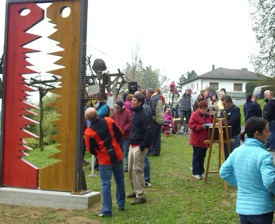 Eine Gruppe von Menschen ist um eine große, farbenfrohe Kunstinstallation in einem Außenbereich versammelt. Einige tragen Jacken und Hosen, andere malen auf Staffeleien. Im Hintergrund sind ein Haus und Bäume zu sehen.