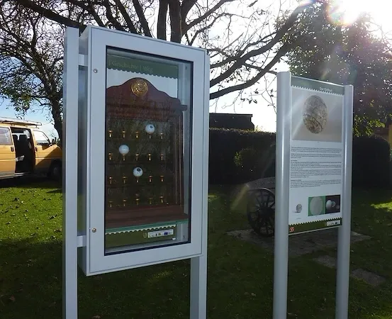 Eine Glasvitrine wird auf einem Rasenfeld im Freien gezeigt, in der Nähe ist ein Lieferwagen geparkt. Die Glasvitrine enthält mehrere Gegenstände, möglicherweise Bälle, und daneben befindet sich ein Schild.