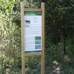 Ein Holzschild mit dem Text 'Fuchs und Hase' steht in einem bewaldeten Gebiet, umgeben von Bäumen und Grün. Es zeigt ein Bild eines Fuchses.