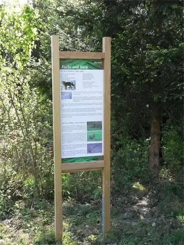 Ein Holzschild mit dem Text 'Fuchs und Hase' steht in einem bewaldeten Gebiet, umgeben von Bäumen und Grün. Es zeigt ein Bild eines Fuchses.