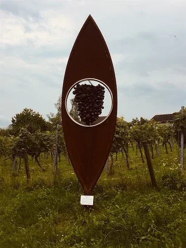 Eine braune Skulptur mit kreisförmigem Ausschnitt steht in einem Weinberg, mit Trauben in der Mitte, unter einem bewölkten Himmel.
