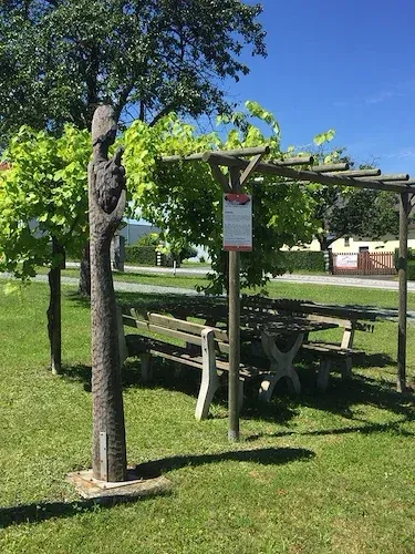 Ein Holzpicknicktisch und eine Bank stehen unter einer Holzpergola in einem Park mit grünen Bäumen und Pflanzen an einem sonnigen Tag.