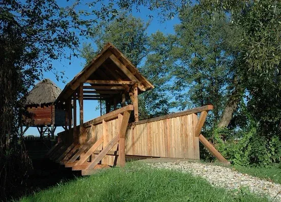 Eine hölzerne Brücke mit einem Strohdach verbindet sich mit einem Holzturm. Umgeben von üppiger Grünfläche, die Szenerie ist vor einem blauen Himmel.