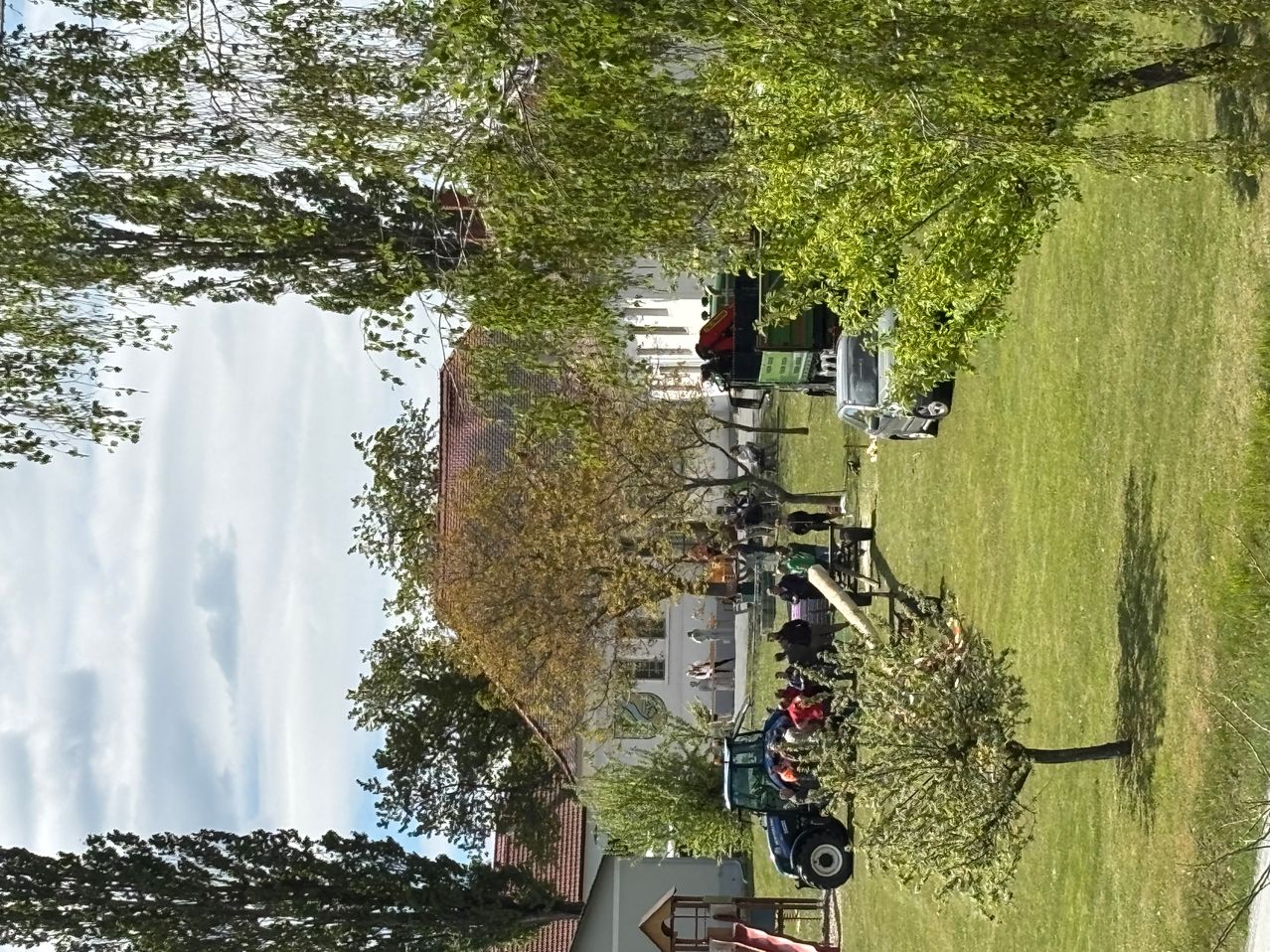 Eine Vogelperspektive eines ländlichen Gebiets mit Bäumen, einem Gebäude und Maschinen. Ein Traktor ist auf einem Rasenfeld geparkt, in der Nähe befinden sich Menschen.