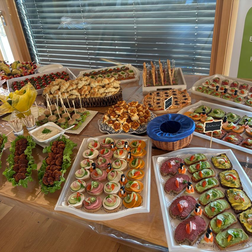 Ein Tisch ist mit einer Vielzahl von Speisen gefüllt, darunter Sandwiches, Salate und Vorspeisen. Der Tisch ist mit einem weißen Tuch bedeckt und die Teller haben Namensschilder. Das Essen ist ordentlich und übersichtlich angeordnet. Der Tisch steht vor einem Fenster mit Jalousien.