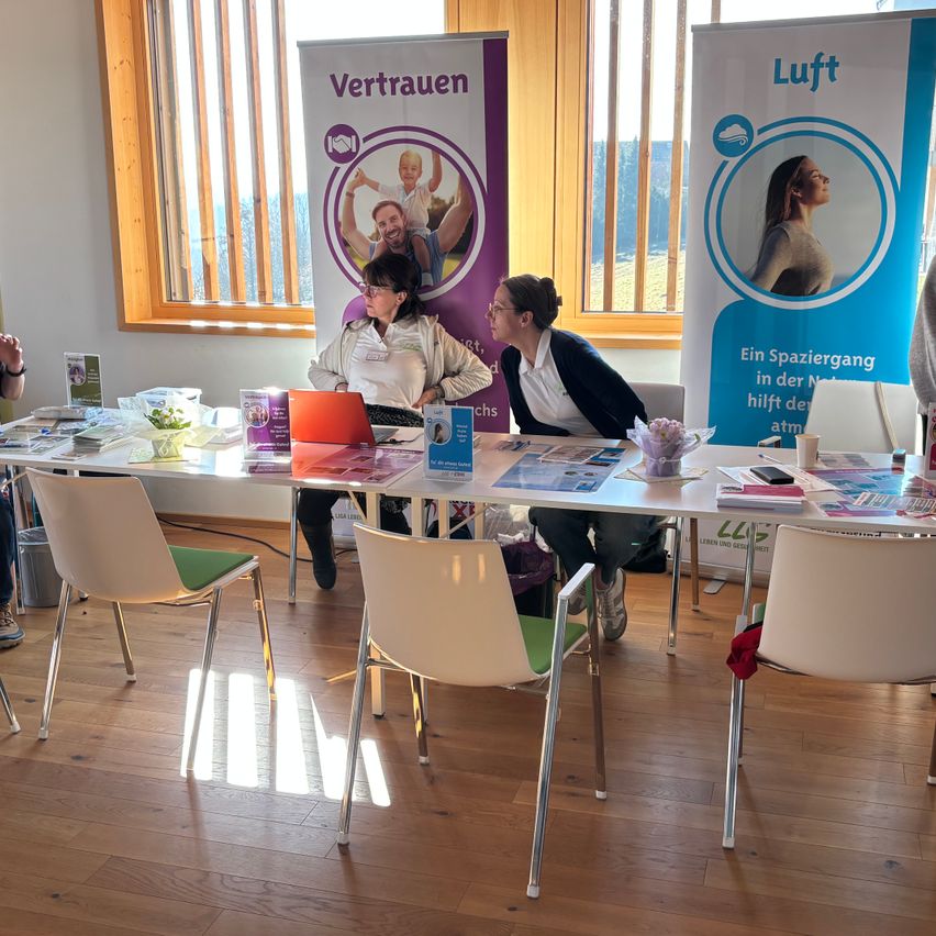 Zwei Frauen sitzen an einem Tisch mit Broschüren und einem Laptop, vor Bannern mit den Worten 'Vertrauen' und 'Luft'. Der Raum hat Holzböden und große Fenster.