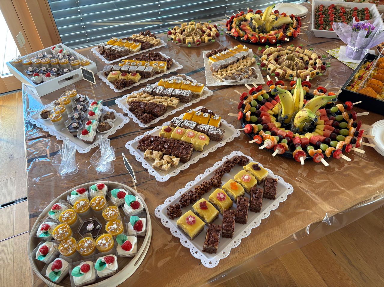 Ein langer Tisch ist mit einer Vielzahl von Desserts bedeckt, darunter Etagenkuchen, Obstspieße und kleine Gebäckstücke. Die Desserts sind auf weißen Tellern mit Spitzenrand angeordnet.