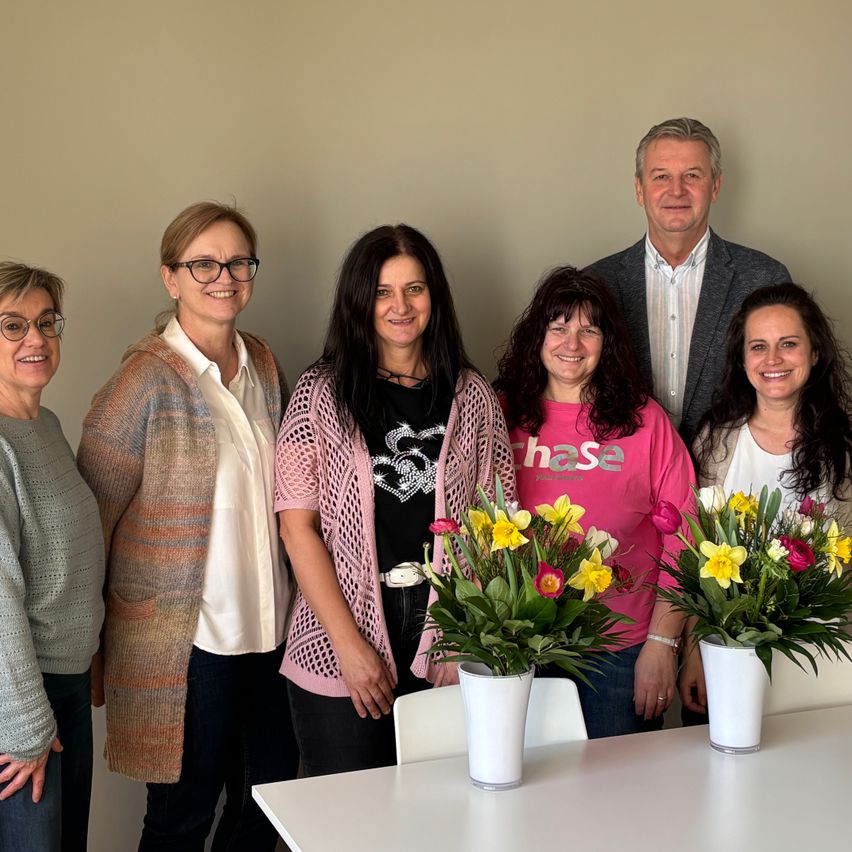 Eine Gruppe von sieben Personen, die lächelnd für ein Foto posiert, mit zwei Blumenvasen auf einem Tisch vor ihnen.