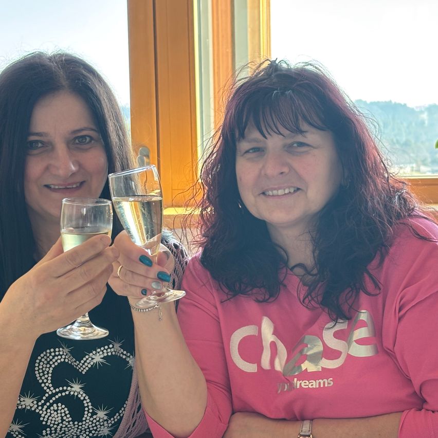 Zwei Frauen sitzen nebeneinander und lächeln, während sie Weingläser halten. Die Frau auf der linken Seite trägt ein schwarzes Oberteil, und die Frau auf der rechten Seite trägt ein rosa Shirt mit weißem Text.