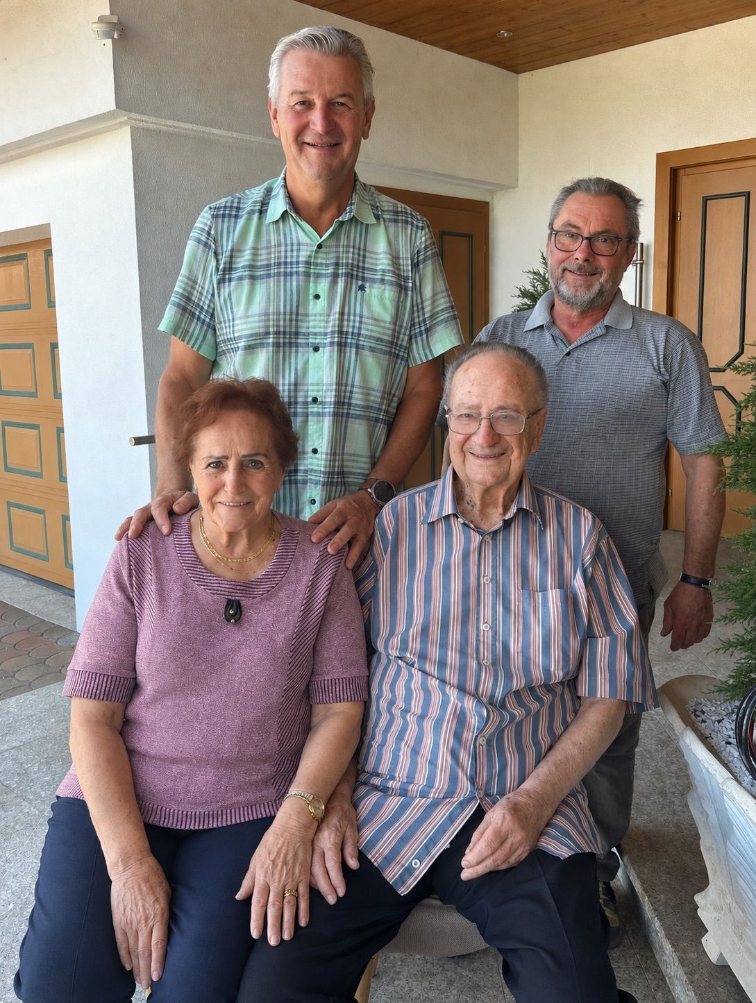 Vier Menschen stehen vor einem Haus. Der Mann auf der linken Seite trägt ein kariertes Hemd. Der Mann auf der rechten Seite trägt eine Brille und eine Uhr. Die Frau in der Mitte trägt eine Halskette. Der Mann neben ihr trägt ein gestreiftes Hemd.
