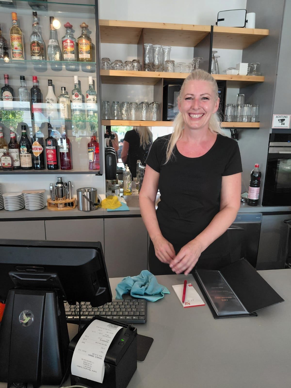 Eine Frau in einem schwarzen T-Shirt steht hinter einer Theke in einer Bar, lächelt und hält einen Stift. Hinter ihr sind Regale mit verschiedenen Likörflaschen zu sehen, und rechts steht eine Mikrowelle.