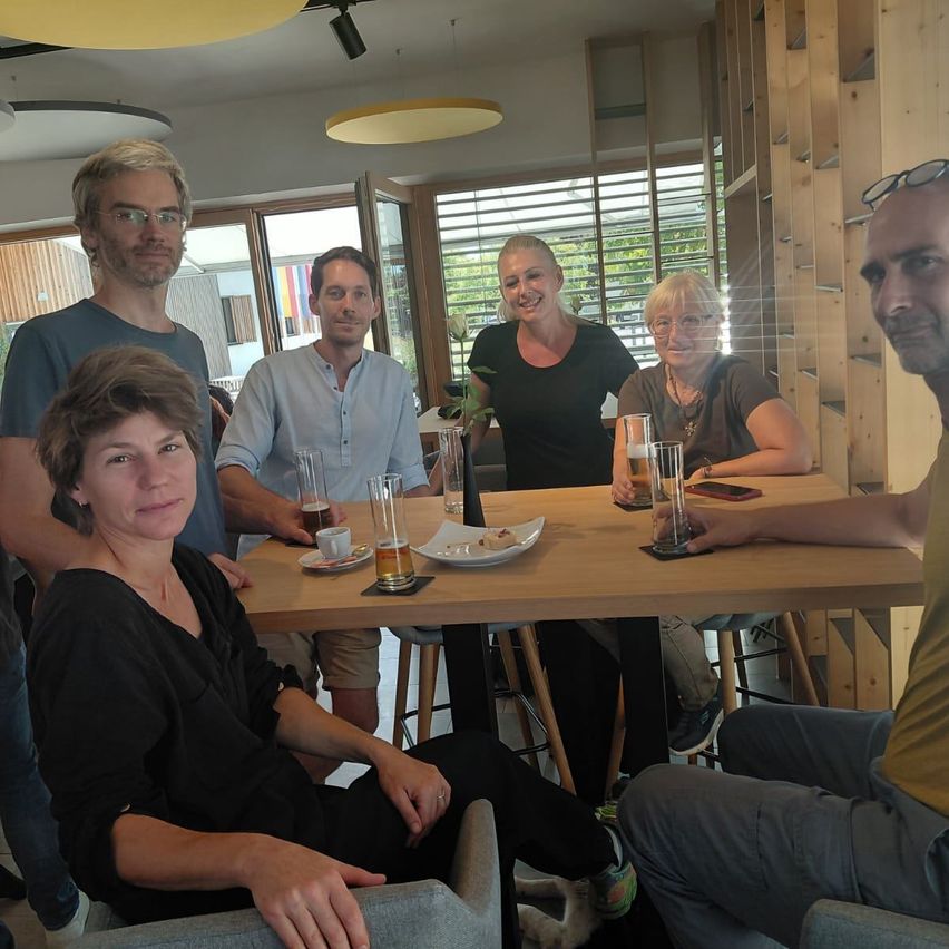 Eine Gruppe von Menschen sitzt um einen Tisch in einem Restaurant, lächelt und posiert für ein Foto. Sie halten Gläser mit Bier und auf dem Tisch steht ein Teller mit einer Tasse.