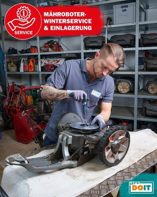A man in a blue shirt is working on a lawn mower in a garage with shelves filled with tools and equipment.