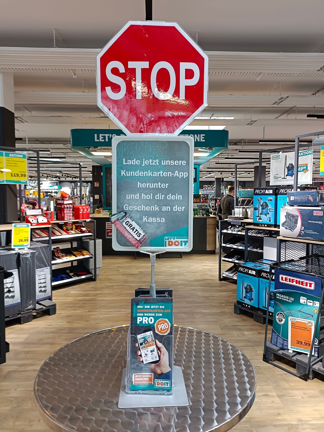 A stop sign above a display reading, 'Load our customer card app now and get your gift at the cash register.' A person holding a phone is shown next to the display.
