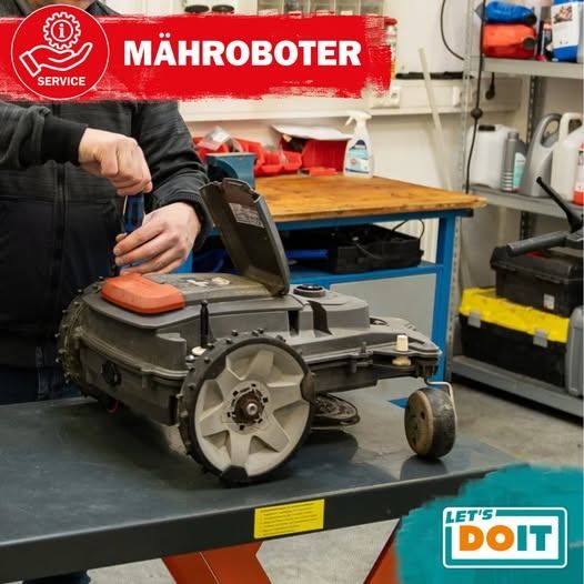 Eine Person arbeitet in einer Werkstatt an einem Roboter-Rasenmäher, umgeben von verschiedenen Werkzeugen und Behältern.