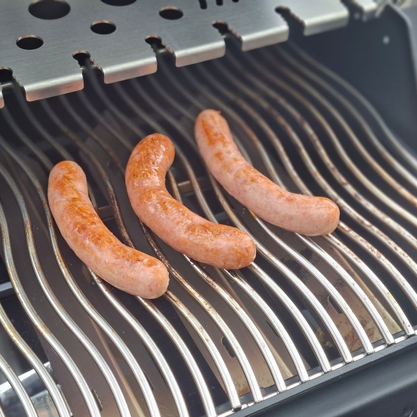 Drei Würste werden auf einem Grill mit einer welligen Muster gekocht. Die Würste sind goldbraun und scheinen gut gegart zu sein.